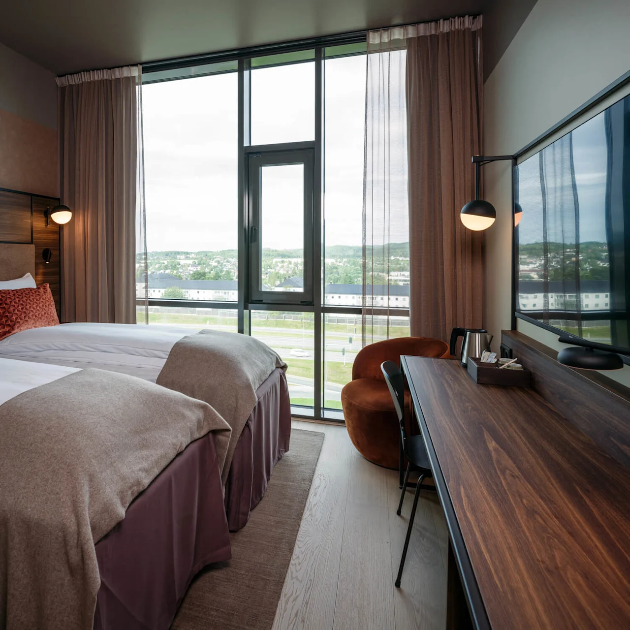 Et moderne hotellrom med to pent oppredde enkeltsenger, pyntet med røde puter, vender mot et stort vindu. Skrivebord i tre med stol og vannkoker ved siden av.