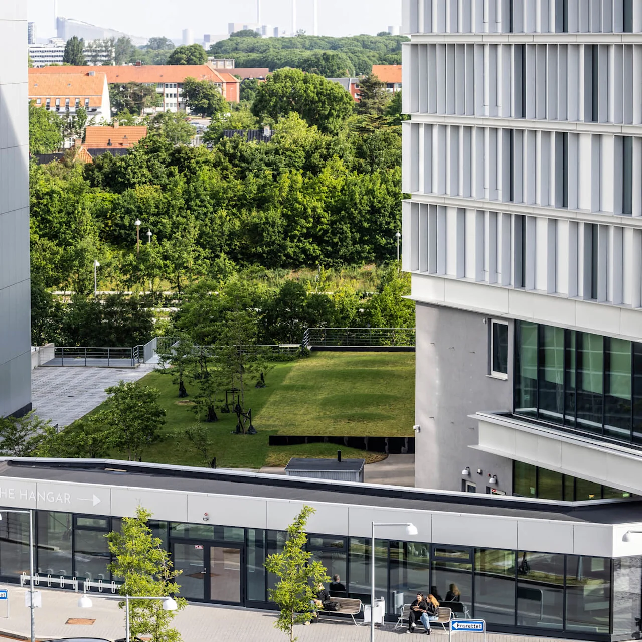 En modern byggnad med eleganta glasfönster står intill frodiga gröna träd och gräs. Skyltar visar "THE HANGAR" med pilar och "Amagerfælled" som antyder en plats.
