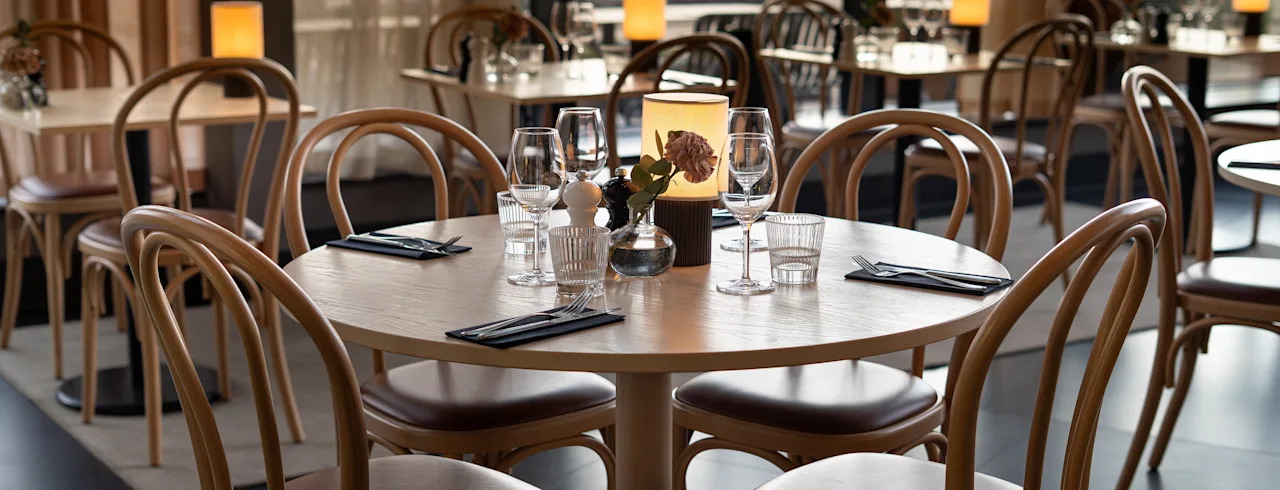Round wooden dining table set with glassware, napkins, and flowers in a dimly lit restaurant with curved chairs and illuminated ambient candles.