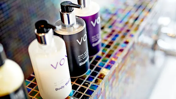 Tre pumpeflasker mærket "vo" (body wash, shampoo, conditioner) står på farverige mosaikfliser ved siden af en vask.