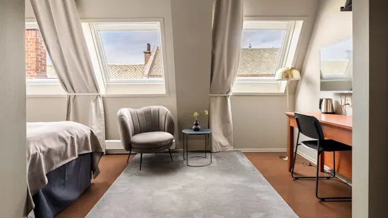 En grå lænestol står ved siden af et lille rundt bord med en vase, nær to tagvinduer. Et skrivebord med stol og lampe fuldender det hyggelige rum.
