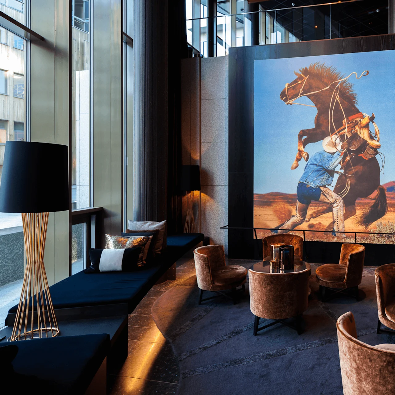En stor, farverig vægmaleri af en cowboy på en stejlende hest pryder et moderne hotellobby med højt til loftet, komfortable siddepladser og udsigt over byen gennem vinduerne.