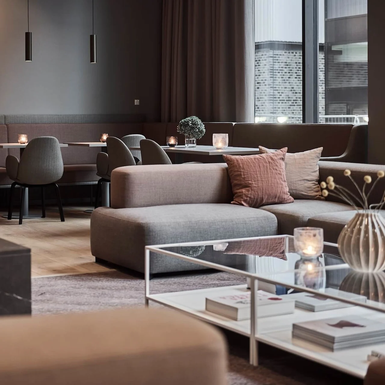 Moderni lounge, jossa on takka, harmaat sohvat ja tyylikäs sohvapöytä. Kynttilät ja minimalistinen sisustus luovat viihtyisän ilmapiirin suurten ikkunoiden taustalla.
