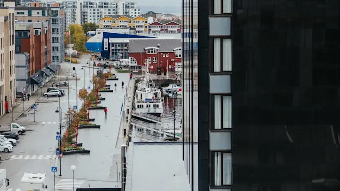 A street lines a harbor with boats docked, surrounded by modern and colorful buildings in an urban waterfront environment.