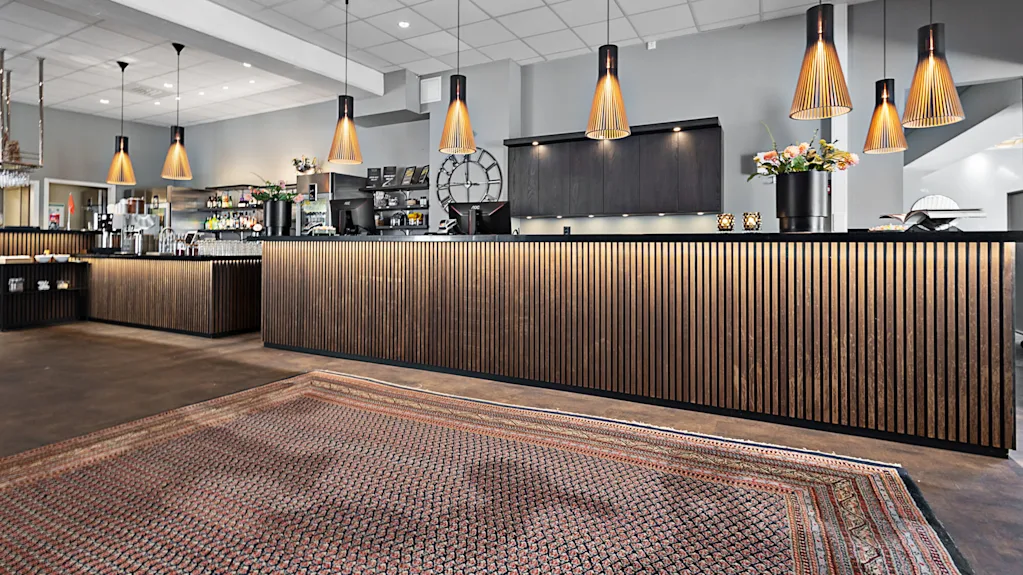 A modern reception desk with wooden panels and pendant lights arranged in a stylish lobby, featuring a patterned carpet and decorative plants on the counter.