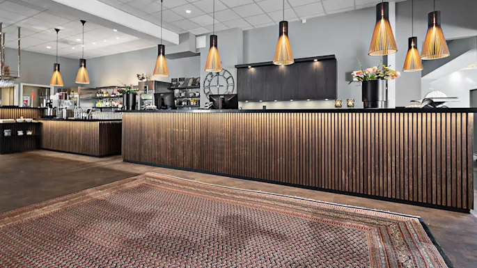 Et moderne resepsjonsbord med trepaneler og hengelampe arrangert i en elegant lobby, med et mønstret teppe og dekorative planter på disken.