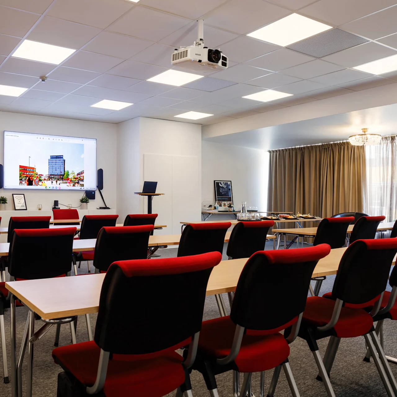 Konferensrum med rader av stolar vända mot en skärm som visar en stadsbild. En projektor är monterad i taket, och bord med förfriskningar finns bak.