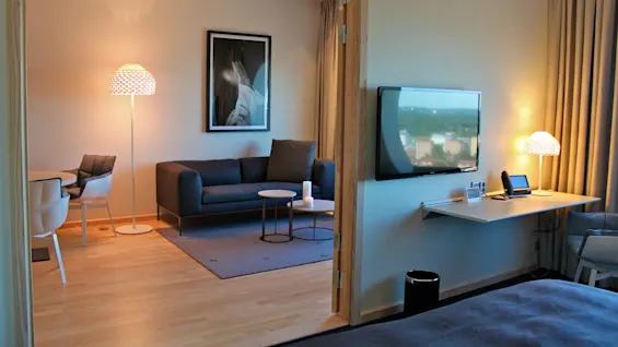 A living room with a dark sofa and small tables sits beside a brightly lit stand lamp and wall art, adjacent to a wall-mounted TV and desk with a lamp.