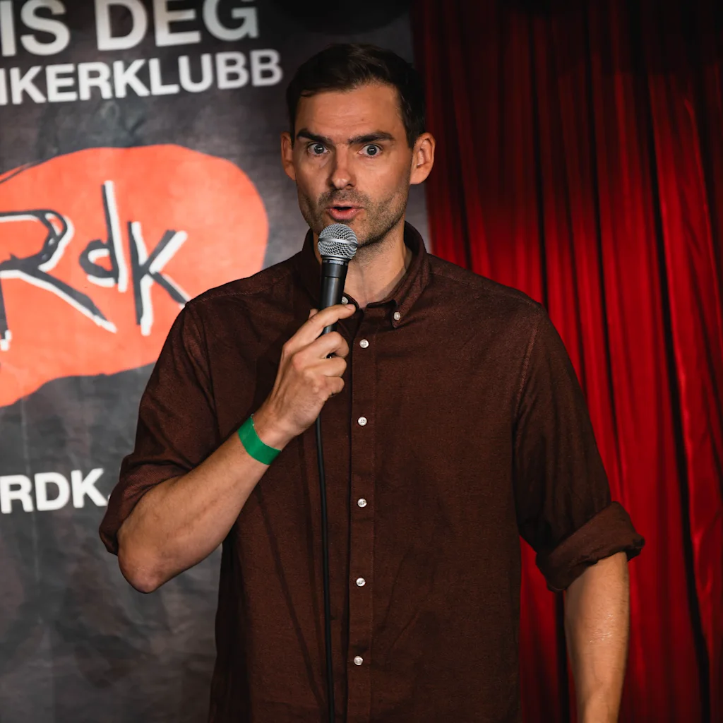 En person opptrer med stand-up komedie, holder en mikrofon. Bak er et rødt sceneteppe og en plakat med teksten REIS DEG KOMIKERKLUBB Rdk RDK.