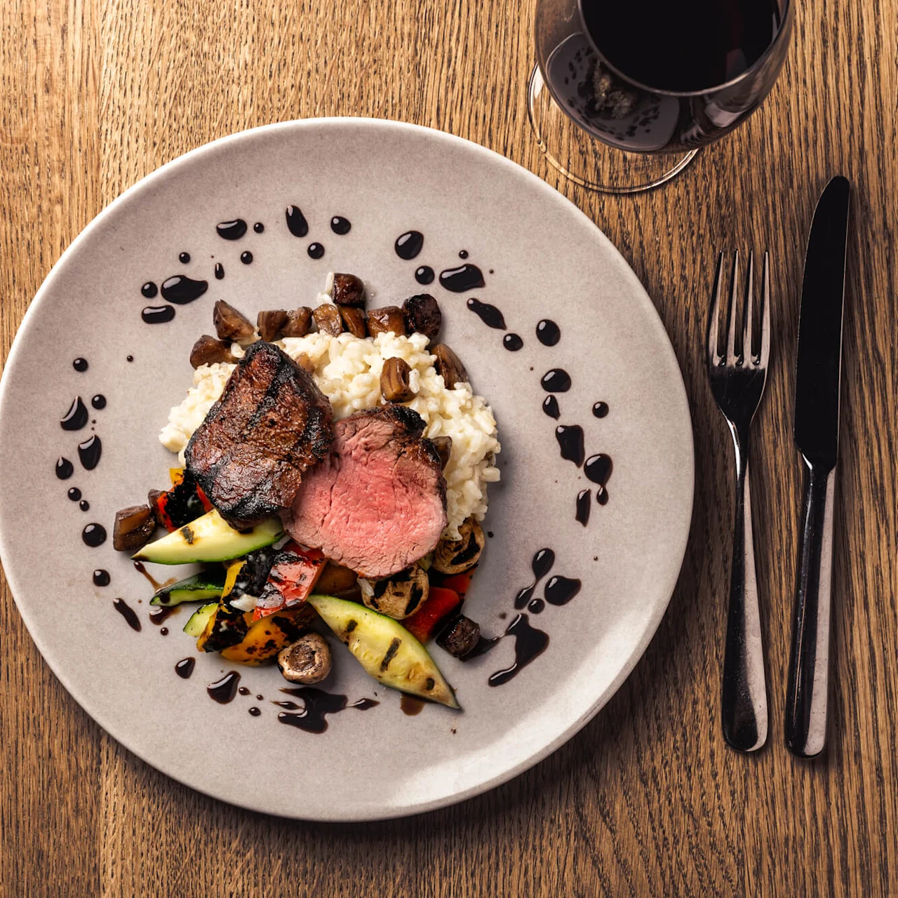 Grilled beef steak on a plate with risotto, vegetables, and sauce droplets sits on a wooden table beside a knife, fork, and glass of red wine.