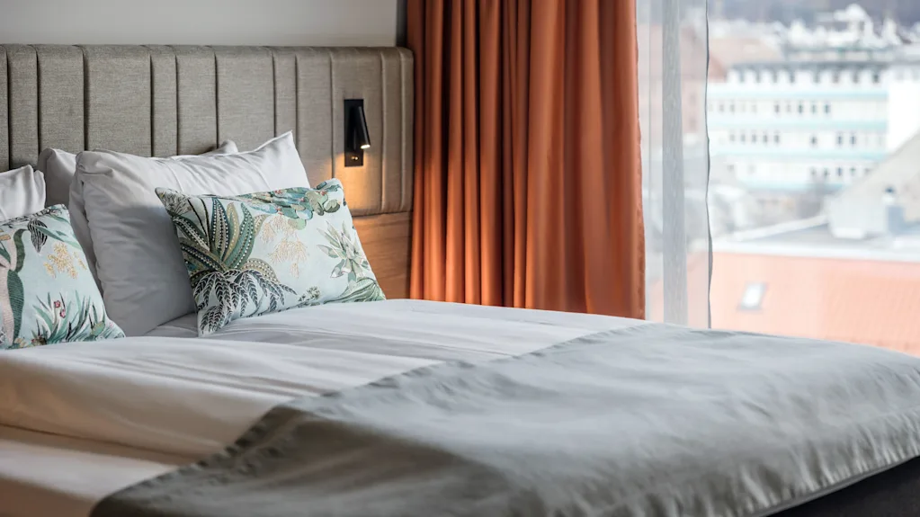 A neatly made bed with patterned pillows rests against a padded headboard, illuminated by a wall lamp, beside orange curtains and a large window overlooking city buildings.
