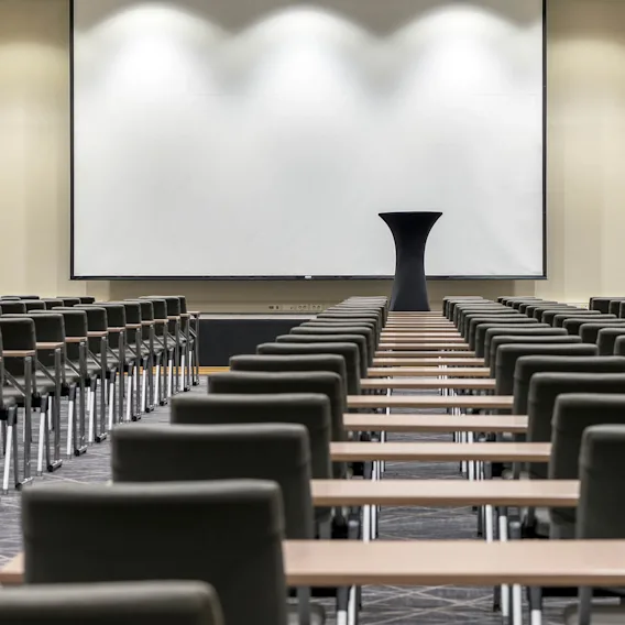 A large conference room features rows of empty chairs facing a white projector screen with a speaker podium in the center. The room is well-lit, with neutral walls and carpeted flooring.