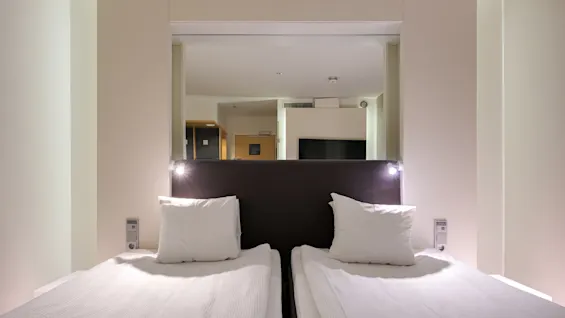 Två prydligt bäddade enkelsängar med vit bäddning, placerade bredvid varandra, är upplysta av små läslampor. En stor spegel speglar ett modernt, neutralt tonat hotellrum.