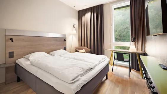 A neatly made bed stands in a hotel room, accented by a large window with brown curtains. A small desk and chair sit nearby, with a lamp providing ambient lighting.