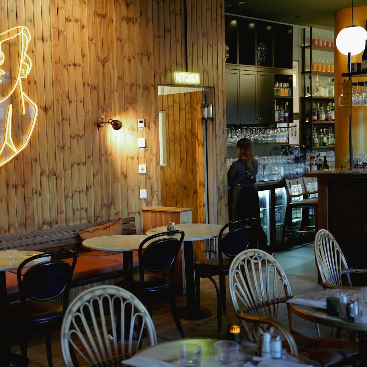 En neon lysprofil af et ansigt på en trævæg med skiltet "KITCHEN", servitrice arbejder i en hyggelig restaurant.