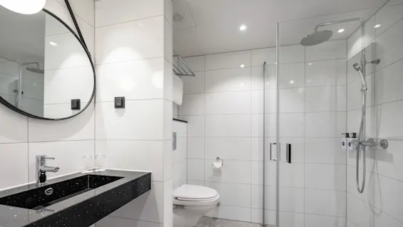Et elegant badeværelse med en sort håndvask og vandhane ved siden af et vægmonteret rundt spejl, med et toilet og en glasomsluttet bruser i nærheden. Hvide fliser og neutrale toner fremhæver den moderne æstetik.