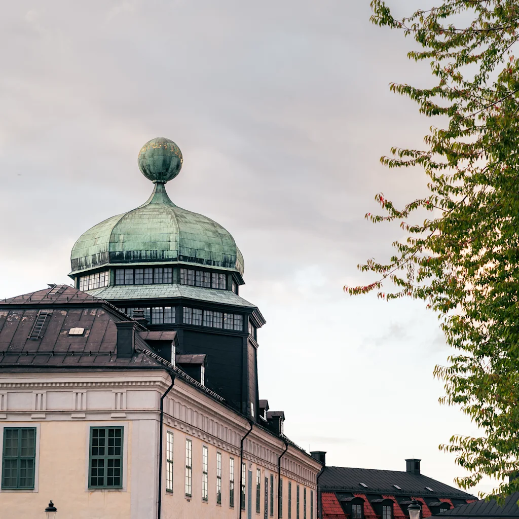 Gustavianumin ikoninen vihreä kupoli Uppsalassa kohoaa pilvisen taivaan alla, vehreiden puunoksien kehystämänä.