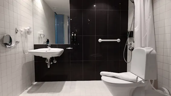 A modern bathroom features a toilet, sink, and mounted showerhead. White and black tiles enhance the contrast. A towel rests on the toilet, with a shower curtain partially open.