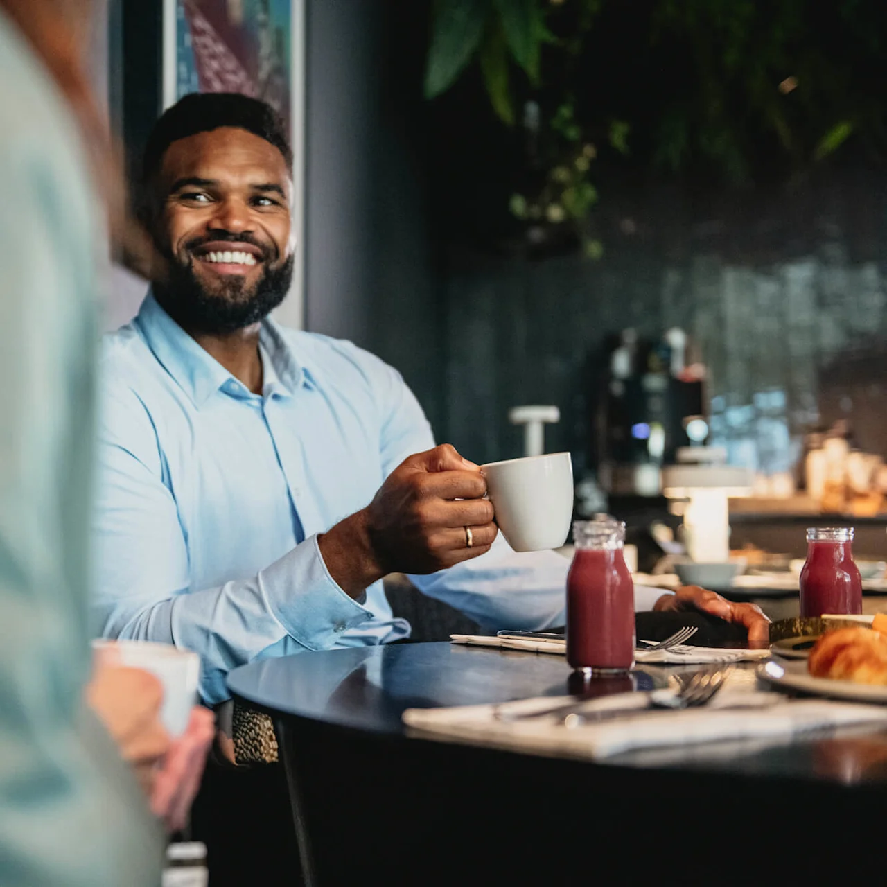 A man in a blue shirt smiles while holding a cup, sitting at a table with juice bottles and pastries in a cozy, dimly lit cafe setting.