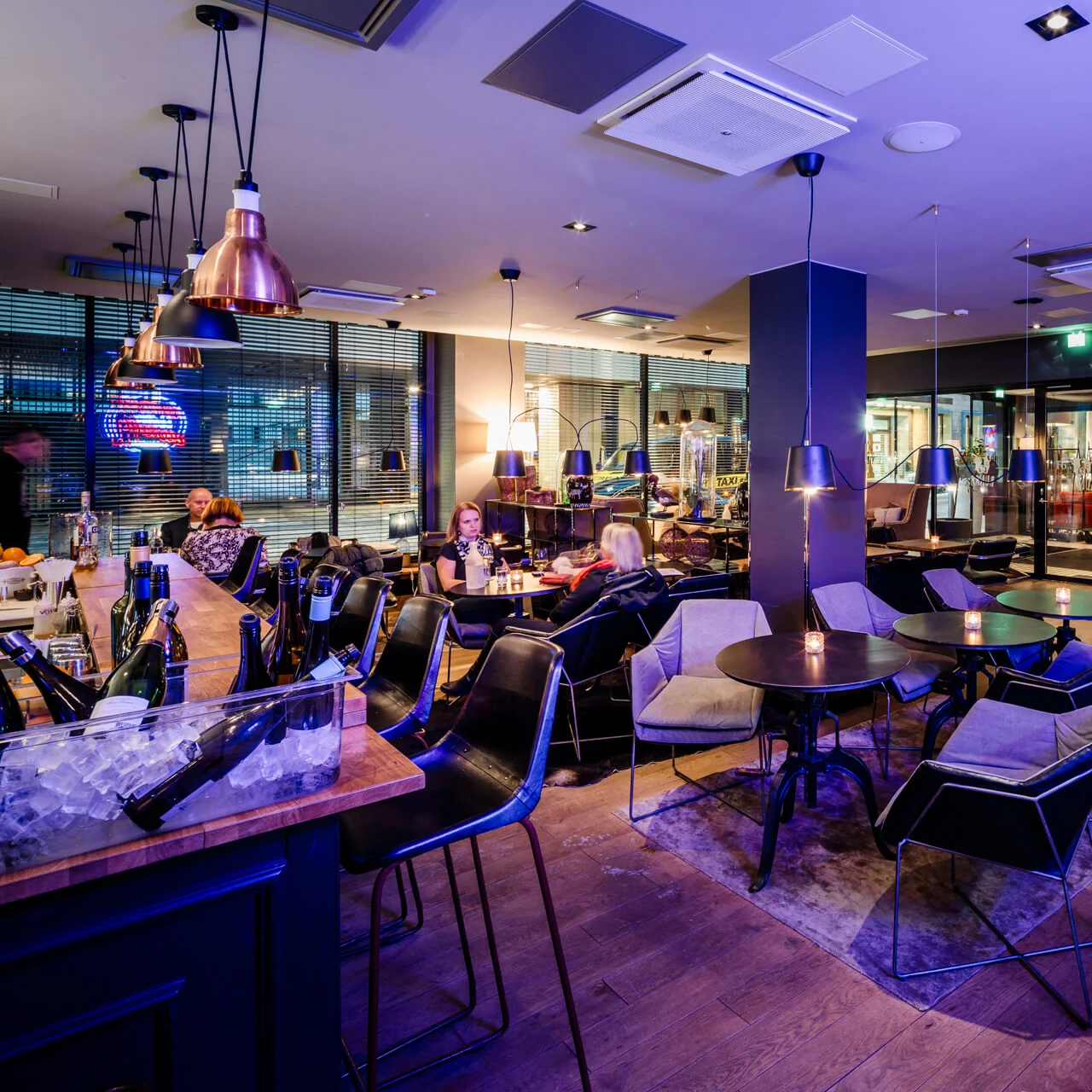 En moderne lounge med svarte bord og stoler, gjester sitter, drikkevarer på baren, hengelamper, og omgivelsesbelysning skaper en varm, avslappet atmosfære.