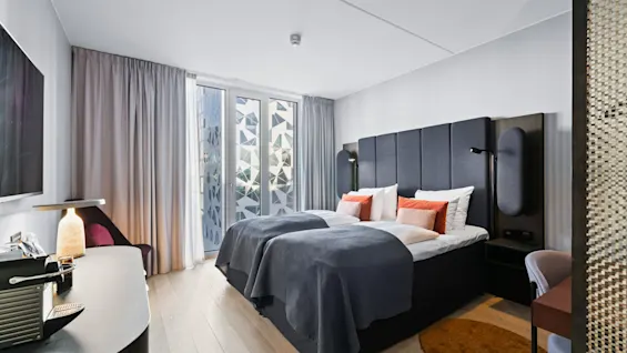 A bed covered with gray blankets and orange pillows is in a modern bedroom. Soft lighting and patterned curtains decorate the room, with furniture including a desk and chair.