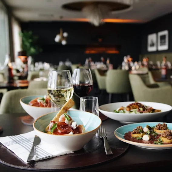 Skålar med gourmetmat och vinglas står på ett träbord i en svagt upplyst restaurang, omgivna av gröna stolar och inramade bilder på mörka väggar.