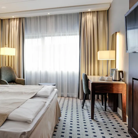 En pent redd dobbeltseng med beige sengetøy står i et godt belyst hotellrom. I nærheten kompletterer et skrivebord med en lampe, stol og vannkoker den koselige, moderne innredningen.
