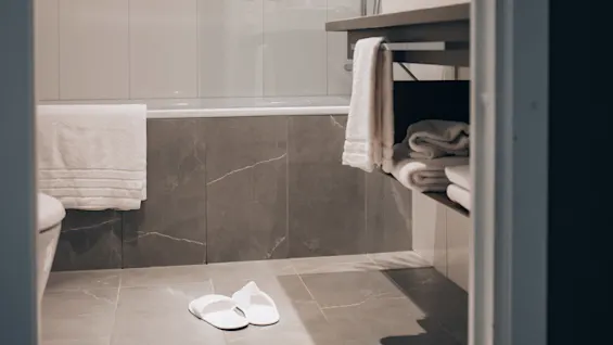 Bathroom with a shower and bathtub. A white towel hangs on the tub, slippers rest on the floor, and folded towels are on a shelf beneath a sink.