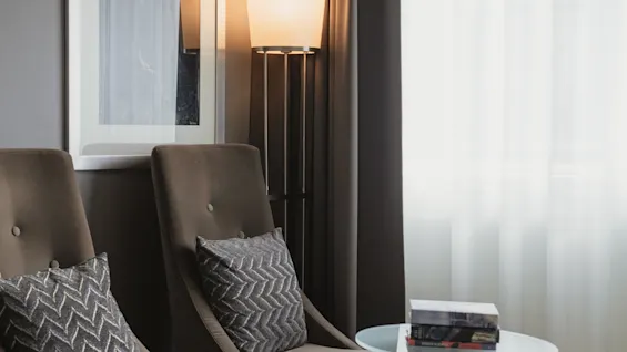 Ett mysigt hotellrumshörn med två fåtöljer, en golvlampa och ett litet bord med böcker vid ett fönster med tunna gardiner.