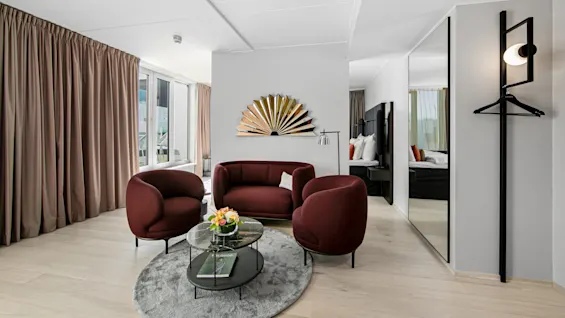 A seating area features three maroon chairs and a sofa around a round coffee table with flowers, set against beige curtains and a decorative wall piece.