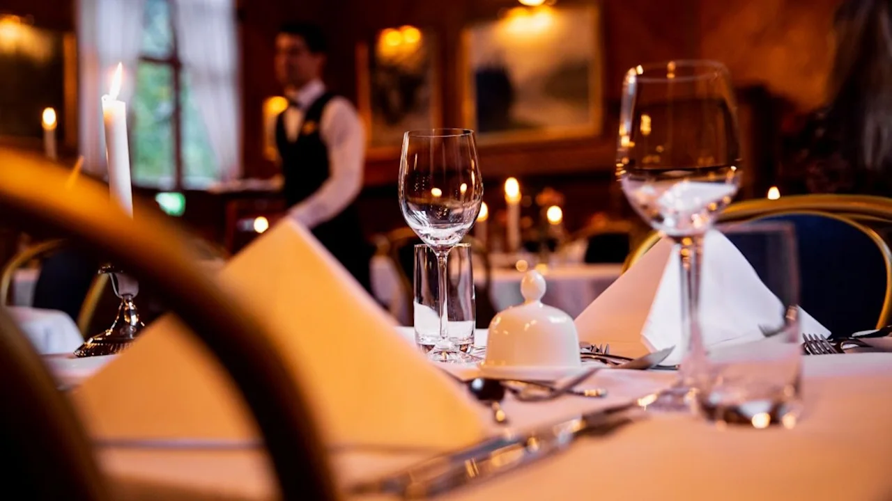 A formally set dining table with white napkins and glasses, surrounded by elegant decor. A waiter in the background walks past, creating an upscale restaurant atmosphere.