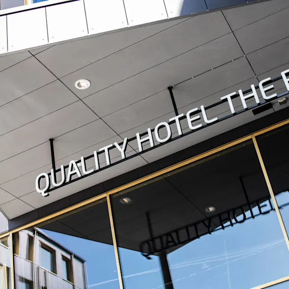 Ett skylt med texten "QUALITY HOTEL THE BOX" hänger på en modern byggnad och reflekteras på stora glasfönster nedanför, vilket antyder en hotellentré i en stadsmiljö.