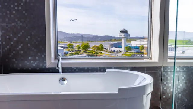 A white bathtub sits inactive in a modern bathroom. Through the window, an aircraft flies above an airport with a control tower surrounded by distant hills.