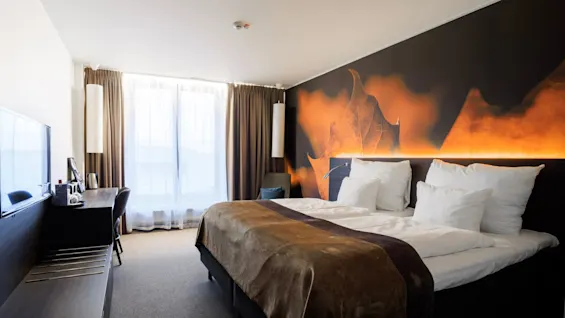 En pænt redt seng med hvide puder og brunt sengetøj, prydet af et stort, oplyst bladmotiv i et moderne hotelværelse med skrivebord og vindue.