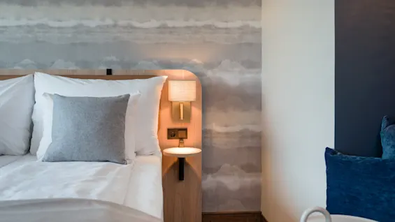 Et koselig hotellrom med seng, nattbordslampe og et komfortabelt sitteområde, med sky-mønstret tapet.