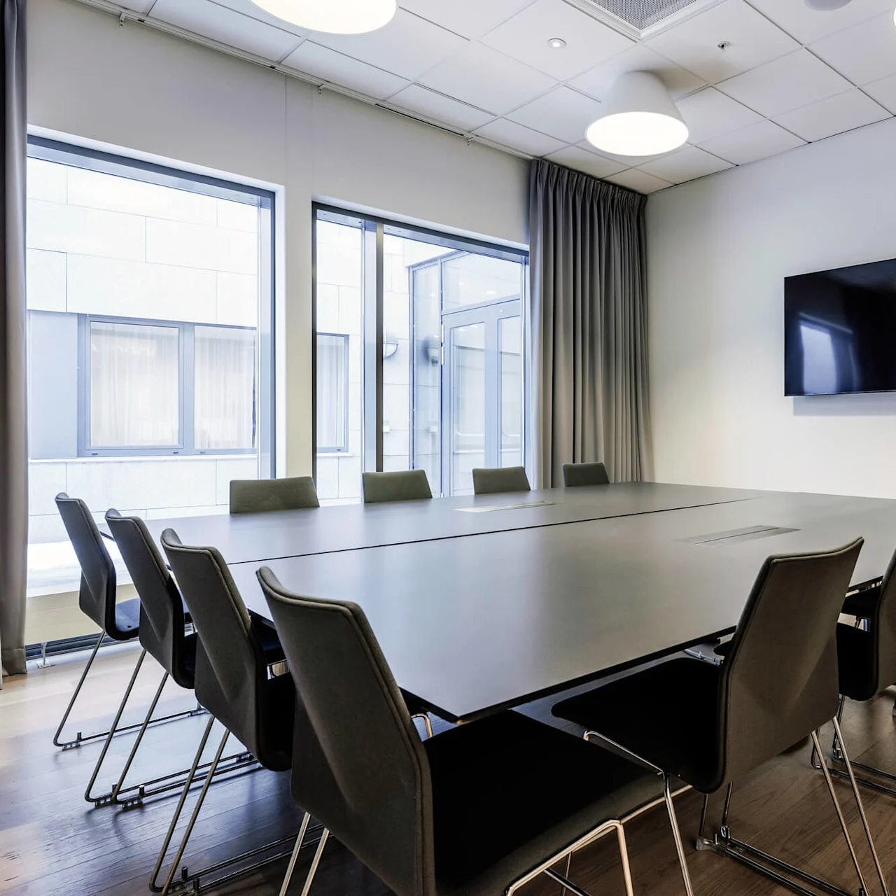 A large conference table surrounded by black chairs sits in a modern office space with large windows, a wall-mounted TV, and wooden flooring. No text present.