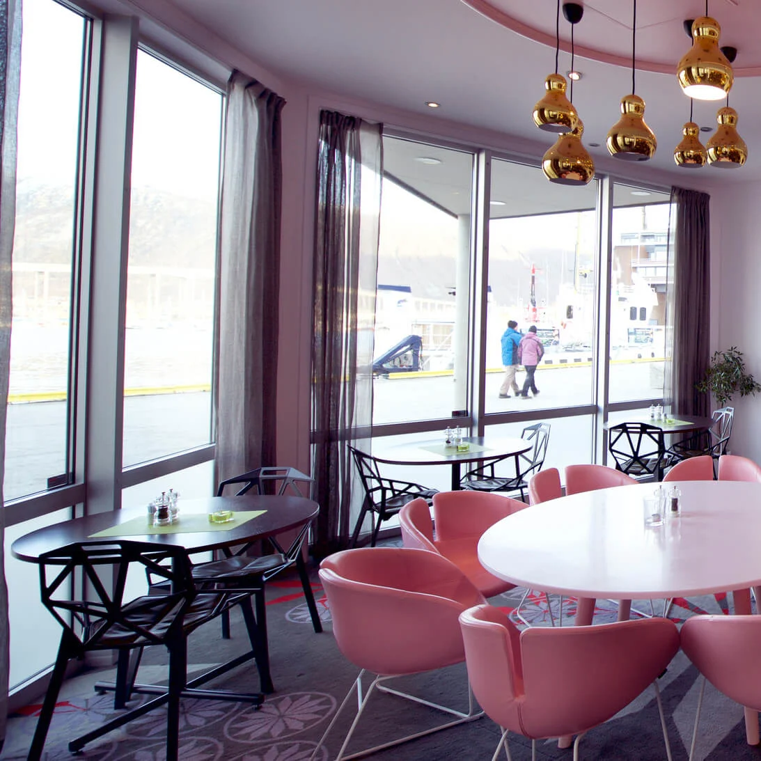 An elegant cafe interior with pink chairs around white tables, large windows, and golden pendant lights.