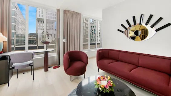A burgundy sofa sits against a wall with an eye-shaped mirror, adjacent to large windows overlooking modern buildings. A desk with a chair and lamp faces outside.
