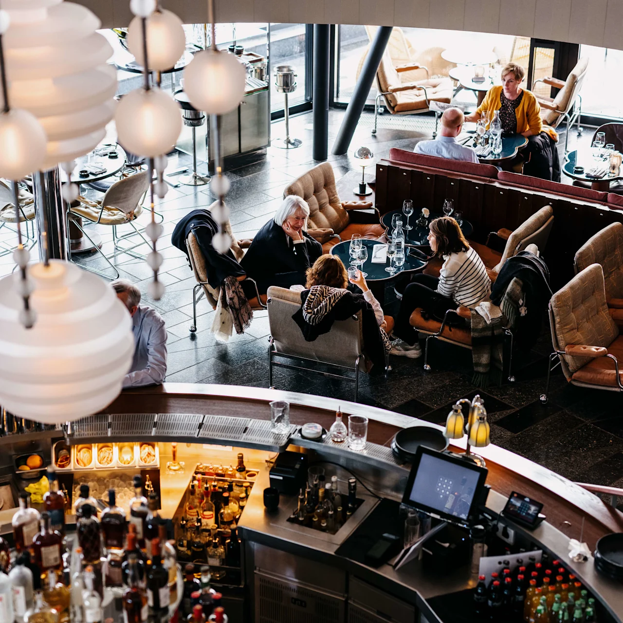 Gæster spiser og socialiserer i en stilfuld, flerniveaurestaurant med hængende globelys, plysmøbler og en dominerende bar.