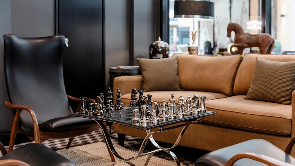 Schackuppsättning på ett elegant bord, med polerade metallpjäser mitt i spelet, omgiven av en beige soffa och en svart läderfåtölj, i ett mysigt, modernt vardagsrum.