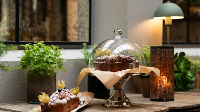 Delicious cakes and pastries displayed on a wooden table with green plants and warm lighting at Home Hotel Grand Bodø.