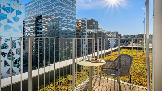 A small round table with a coffee cup and croissants sits on a balcony with a wicker chair, overlooking modern buildings under a bright sun in an urban setting.