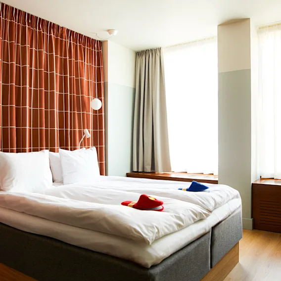 Viihtyisä hotellihuone, jossa on suuri sänky, valkoiset liinavaatteet ja tossut, kahden suuren ikkunan luonnonvalossa.