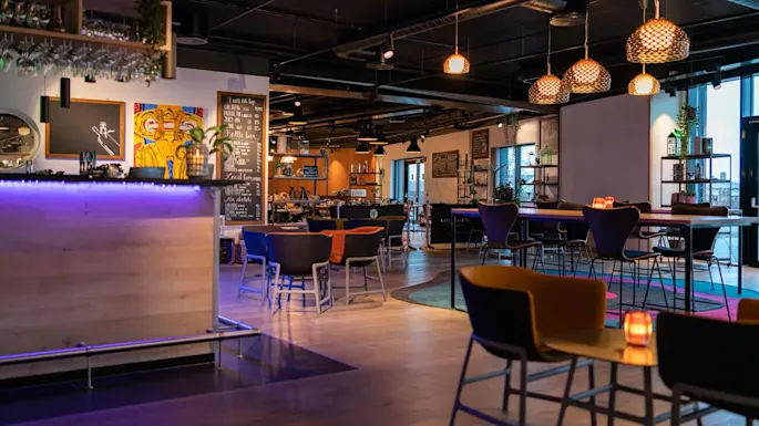 A cozy bar interior with a wooden counter lit by blue lights and modern pendant lamps, surrounded by tables and chairs. Chalkboards display menus on the walls.