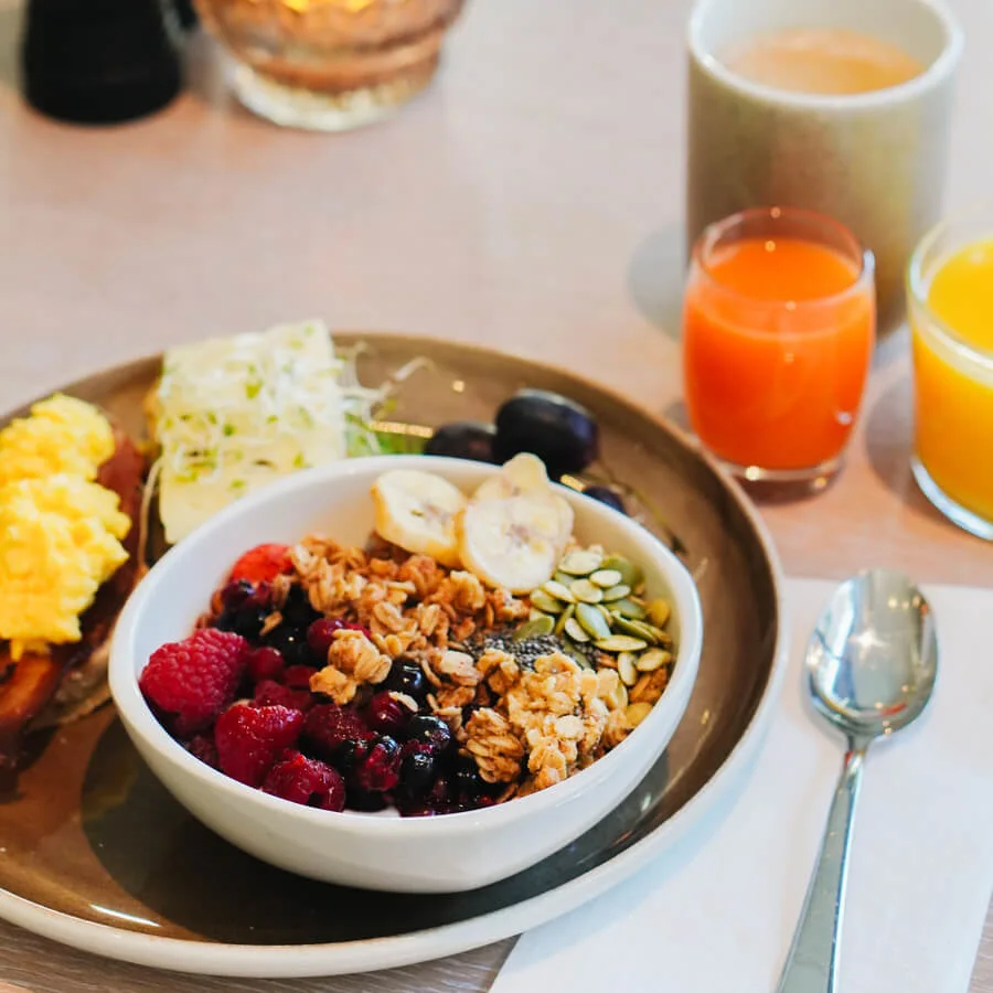 A bowl of granola with fruits sits on a table, surrounded by glasses of juice, a candle, and silverware, creating a cozy breakfast setting.