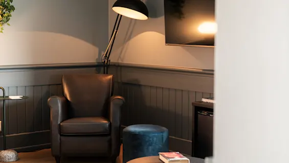 En lænestol står i hjørnet, oplyst af en høj lampe. I nærheden er et lille bord med bøger og et fjernsyn hænger på væggen i et hyggeligt rum.
