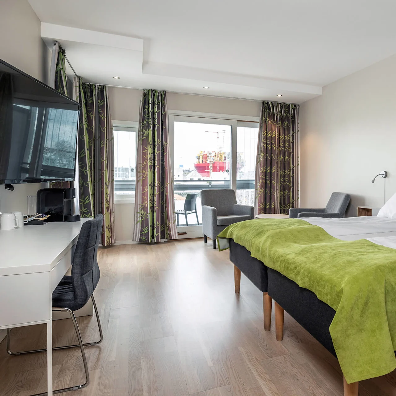 Ett prydligt hotellrum med en dubbelsäng täckt med en grön pläd, vänd mot en TV och ett skrivbord; stora fönster med mönstrade gardiner erbjuder utsikt över en hamn.