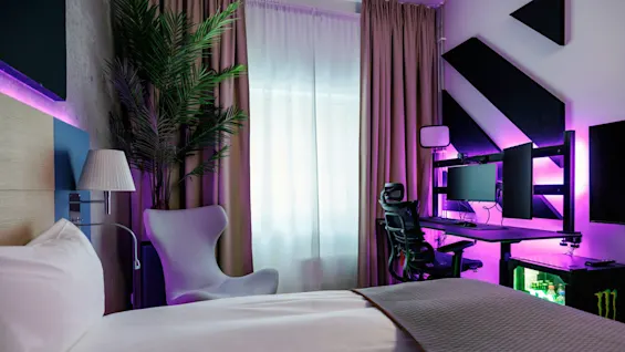 A bedroom features a neatly made bed, a sleek desk with dual monitors emitting purple light, an ergonomic chair, and a modern lamp, under softly draped curtains beside a large plant.