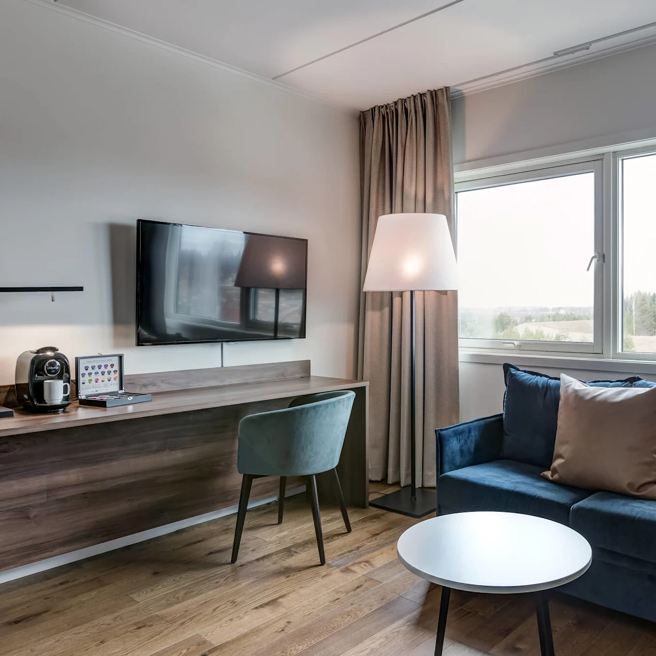 A cozy room features a blue sofa with a brown cushion and a round coffee table, positioned next to a window showing a landscape view. Nearby is a desk with a TV, coffee machine, and a chair.