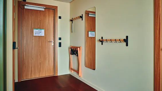 En hotellromsinngang med tredør, speil, kleskroker og bagasjestativ, klar for en gjests ankomst.